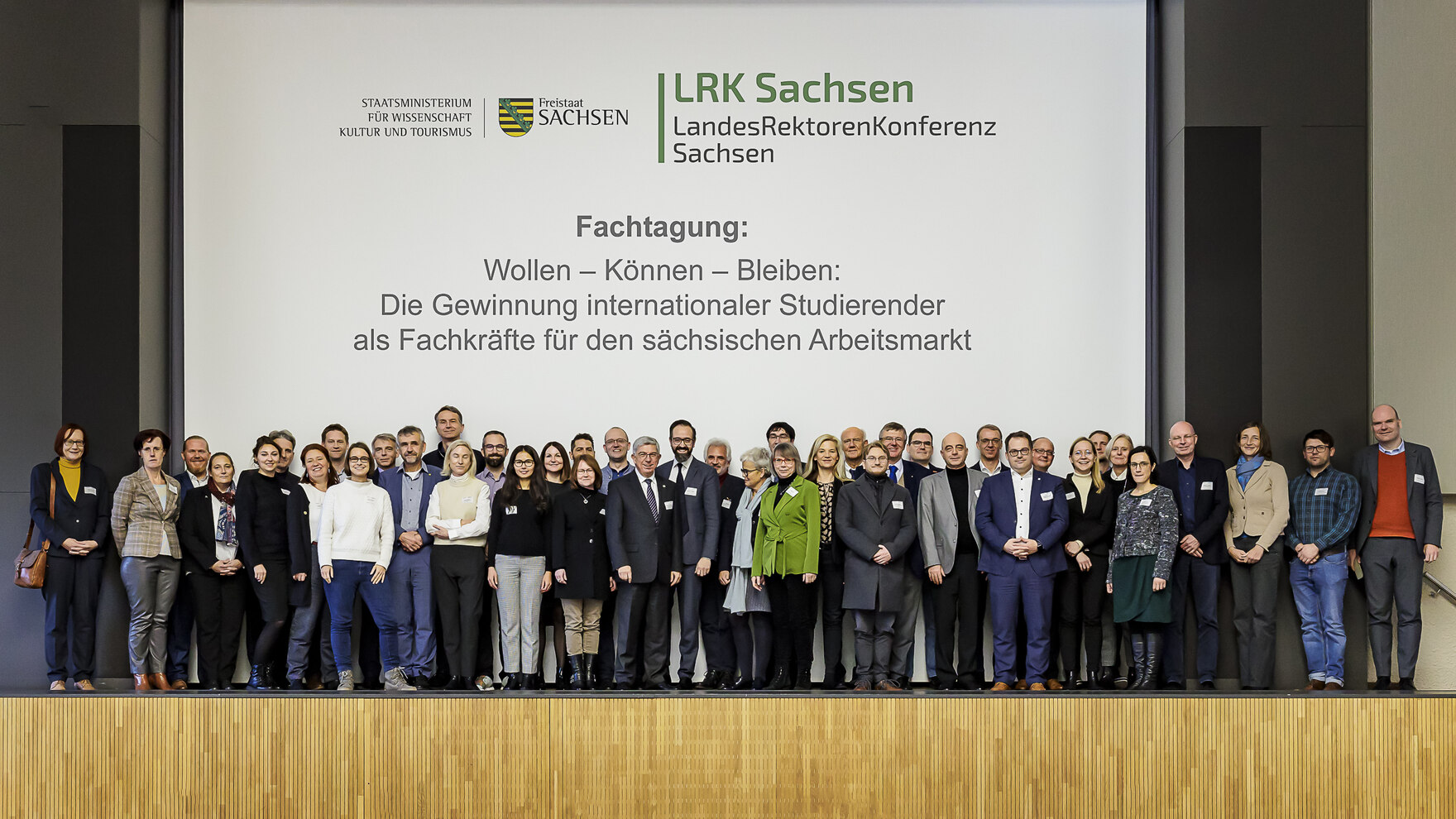 Gruppenbild von der Fachtagung mit Schrift im Hintergrund: Wollen - Können - Bleiben: Die Gewinnung internationaler Studierender als Fachkräfte für den sächsischen Arbietsmarkt. Darüber Logos des SMWK und der LRK.